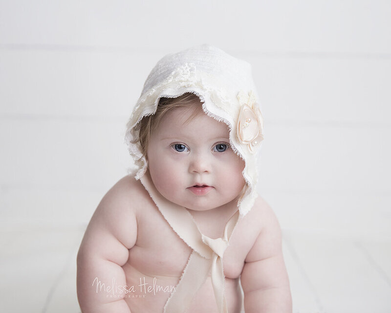 Girl sits with white bonnet and  pink flower for her first birthday portraits.