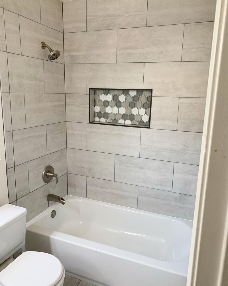 A newly tiled tub shower combo with horizontal large grey tiles and a shampoo box tiled with small grey and white hexagon tiles.