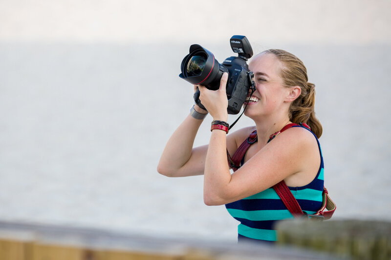 Jen Stitt holding a super wide-angle lens and Profoto gear on a beach during an engagement session, showcasing her professional setup and dedication to capturing stunning and creative oceanfront moments.