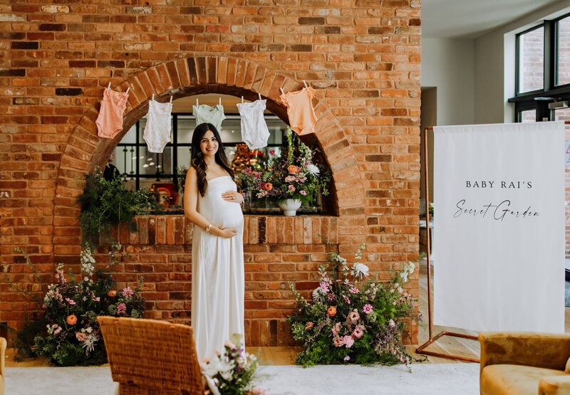 Mom-to-be posing at a baby shower with floral décor, hanging baby clothes, and a custom sign reading ‘Baby Kai’s Secret Garden,’ styled by Yours Truly, a wedding planner and designer in New Jersey