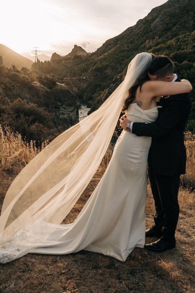 a bride hugs her husband in front of a dramatic queenstown new zealand landscape at sunset