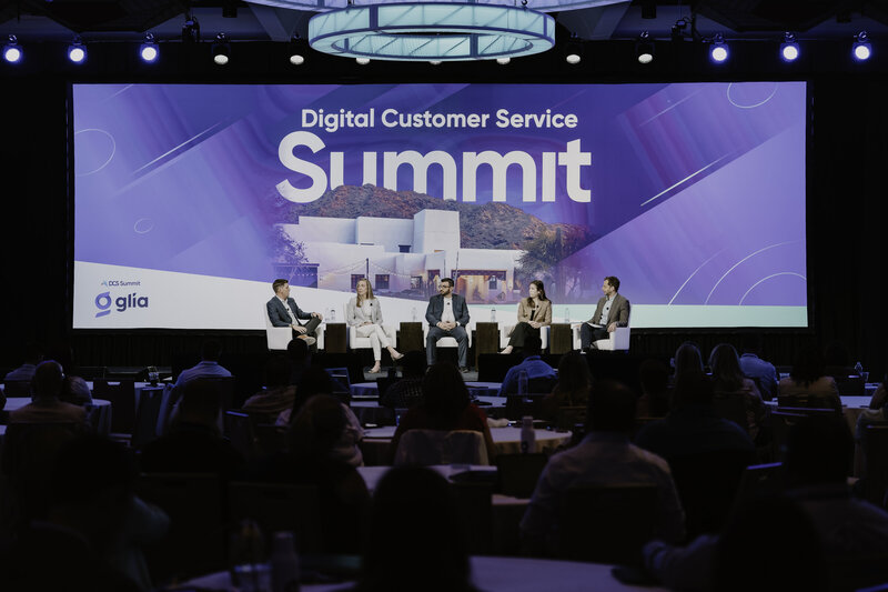 Four panelists at the Digital Customer Service Summit are engaged in discussion on stage, each seated in front of a large backdrop with the event title and an image of mountainous landscape. The audience, viewed from behind, focuses on the speakers. The setting features modern conference lighting and a formal yet inviting atmosphere.