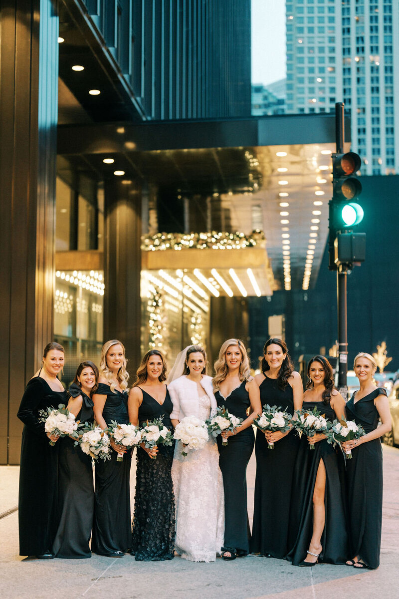 Bride and her bridesmaids dressed in sleek black gowns, holding silver bouquets, standing in front of a modern building with sharp lighting at The Langham Chicago.