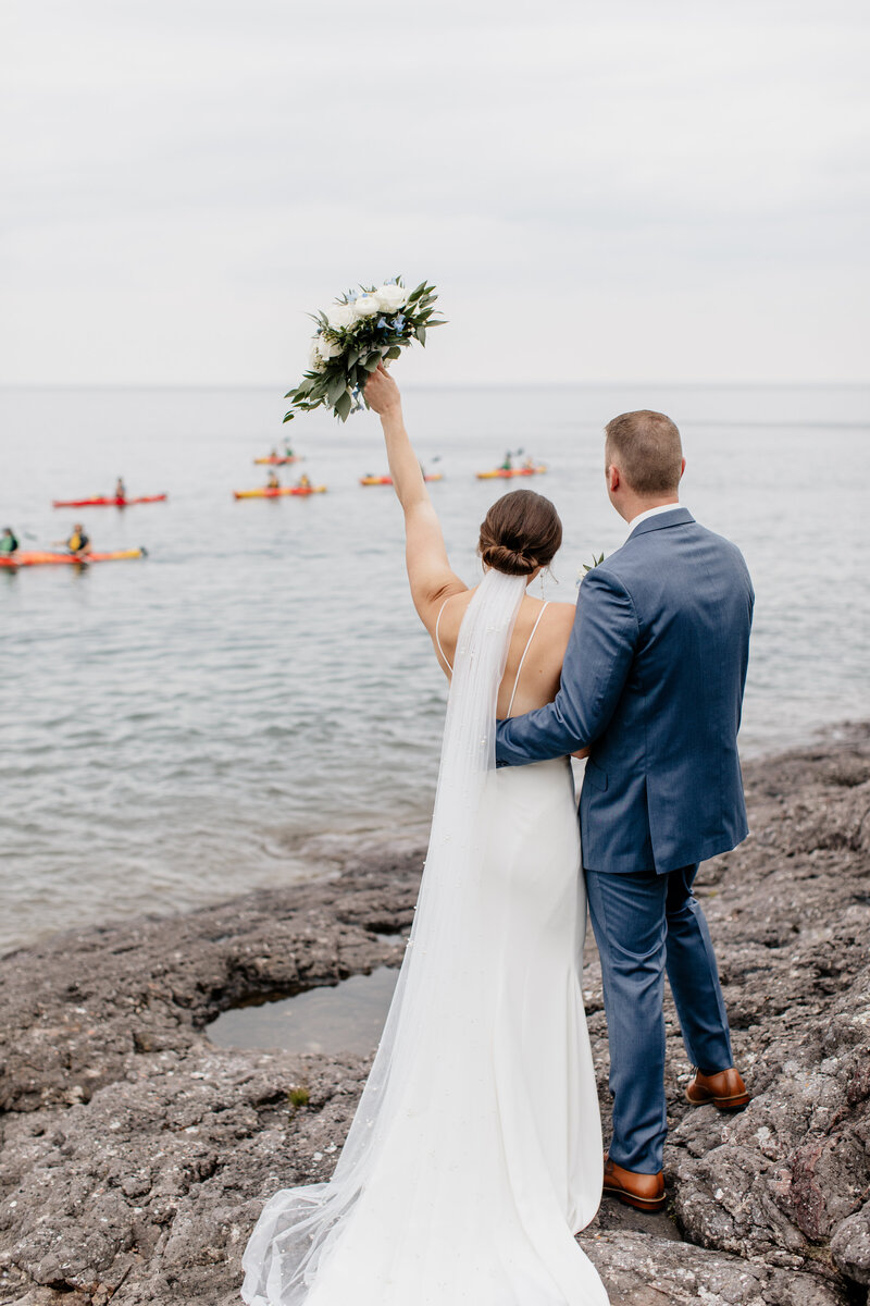 Elopement ceremony at Three Points Lodge in Tofte, MN, with the bride cheering and holding her bouquet as a group of kayakers pass by on Lake Superior, celebrating the couple, captured by a wedding photographer in MN.