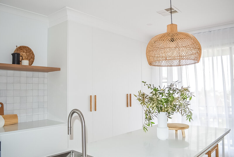 Minimalist kitchen with white cabinets, a woven pendant light, and a vase with green branches on the counter—an example of luxury home design Newcastle appreciates.