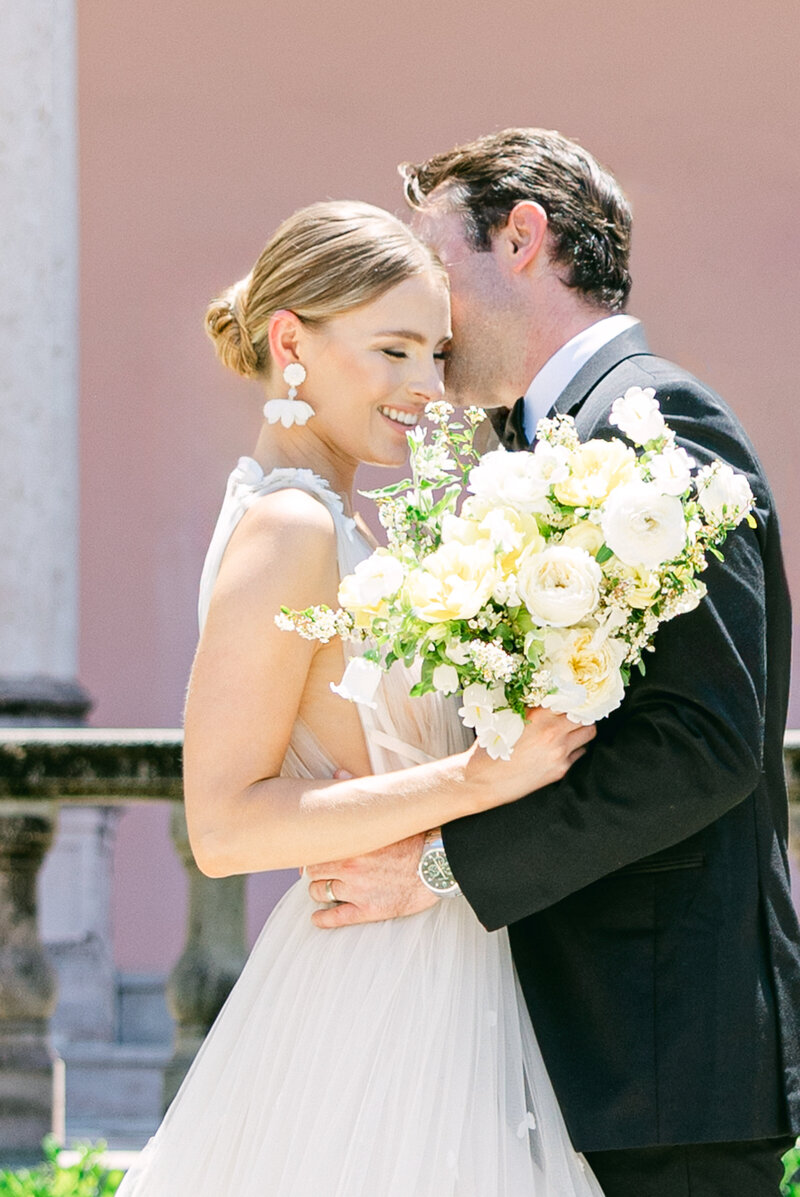 A joyful bride embraces her groom while holding a lush white and yellow floral bouquet at The Ringling Museum in Sarasota, Florida. Captured by Amia Marcell destination wedding Photographer. 