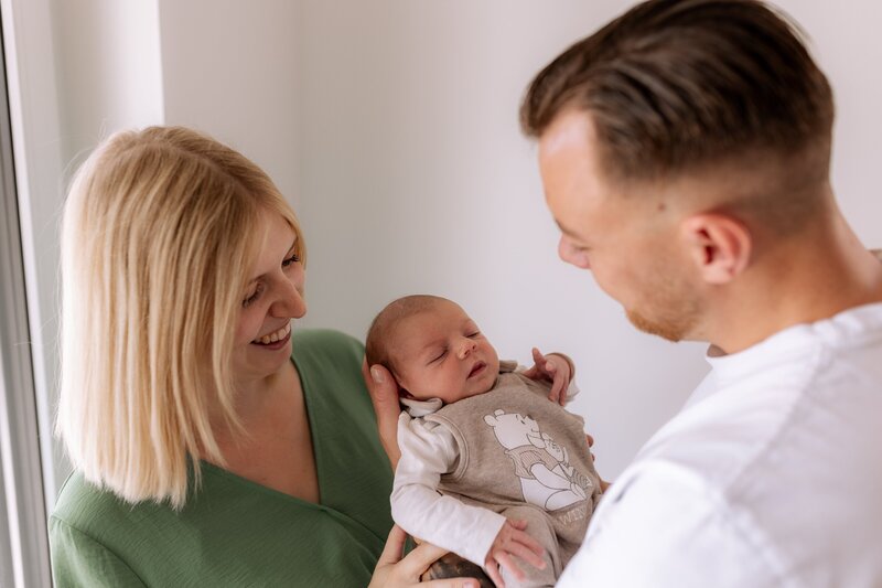 Vater haelt Baby vor sich und Mutter steht daneben und lacht