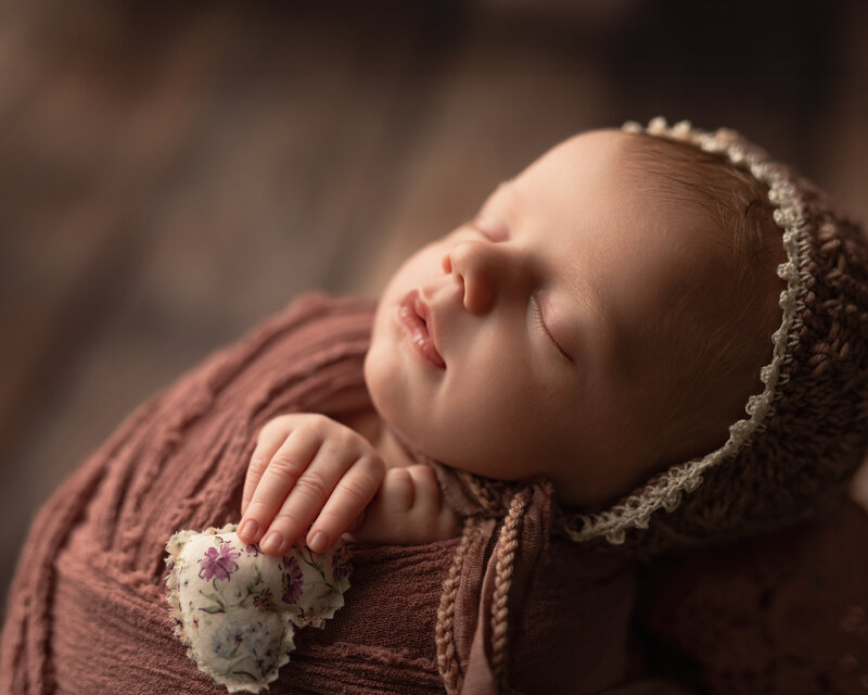 This heartwarming close-up captures every tender detail of a sleepy newborn girl in dusty rose, holding a handcrafted heart.