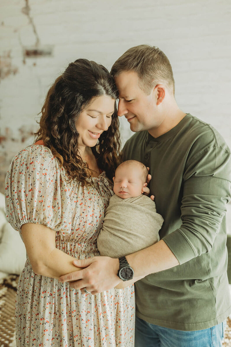 mom in a floral dress and dad in a green henley holding their newborn boy with their eyes closed