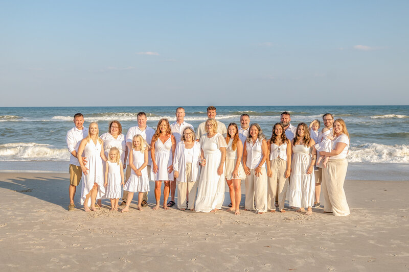 A large family stands together on a sunny beach, dressed in coordinated white outfits. The bright ocean waves provide a lively backdrop, perfect for artistic family photography in Wilmington, NC, capturing vibrant and joyful family moments.