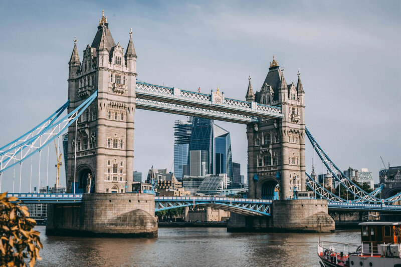 Tower Bridge in London spans the River Thames on a clear day. The iconic structure's two towers and bascule walkways are visible, with modern skyscrapers in the background. A small boat is on the river in the foreground, perhaps part of a journey curated by a European travel specialist.