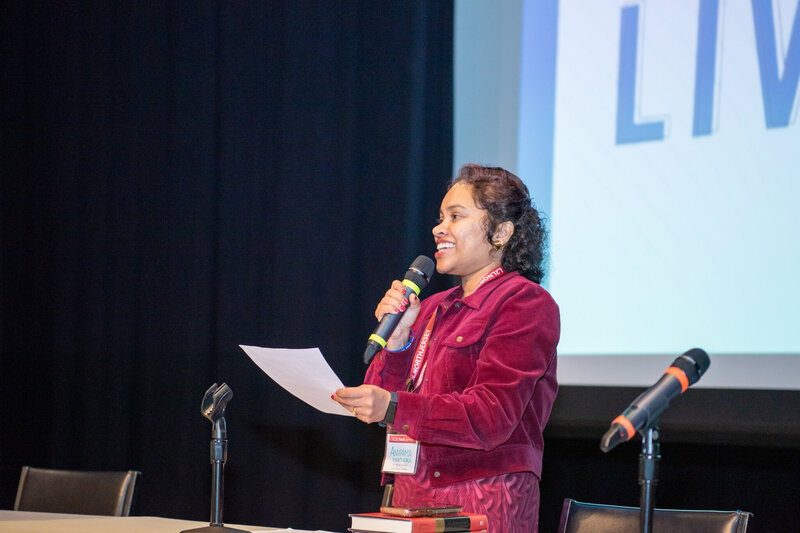 The female host of the Live2Lead conference talking to the audience on a stage holding a microphone.