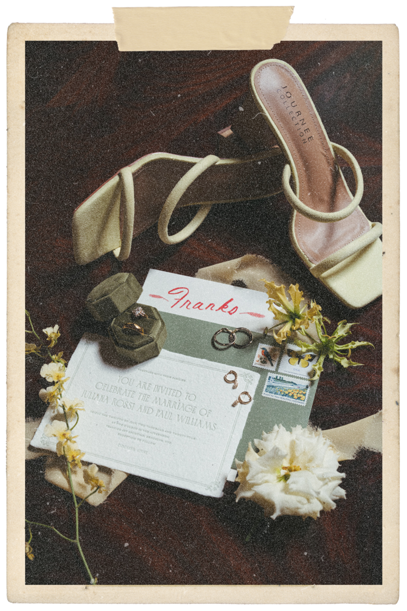 Aged photo of flatlay with green heels, an invitation suite, flowers and rings on a dark wood floor. A piece of tape at the top of the photo.
