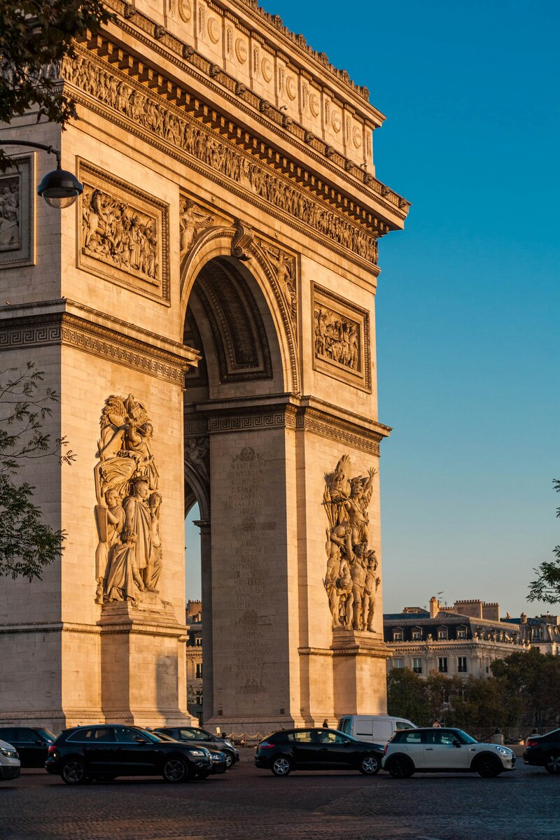 A close-up of the Arc de Triomphe in Paris, France, bathed in golden light against a clear blue sky. Explore bespoke luxury travel experiences, private guided tours, and exclusive Parisian escapes curated by Nadia Forte Travel & Events.