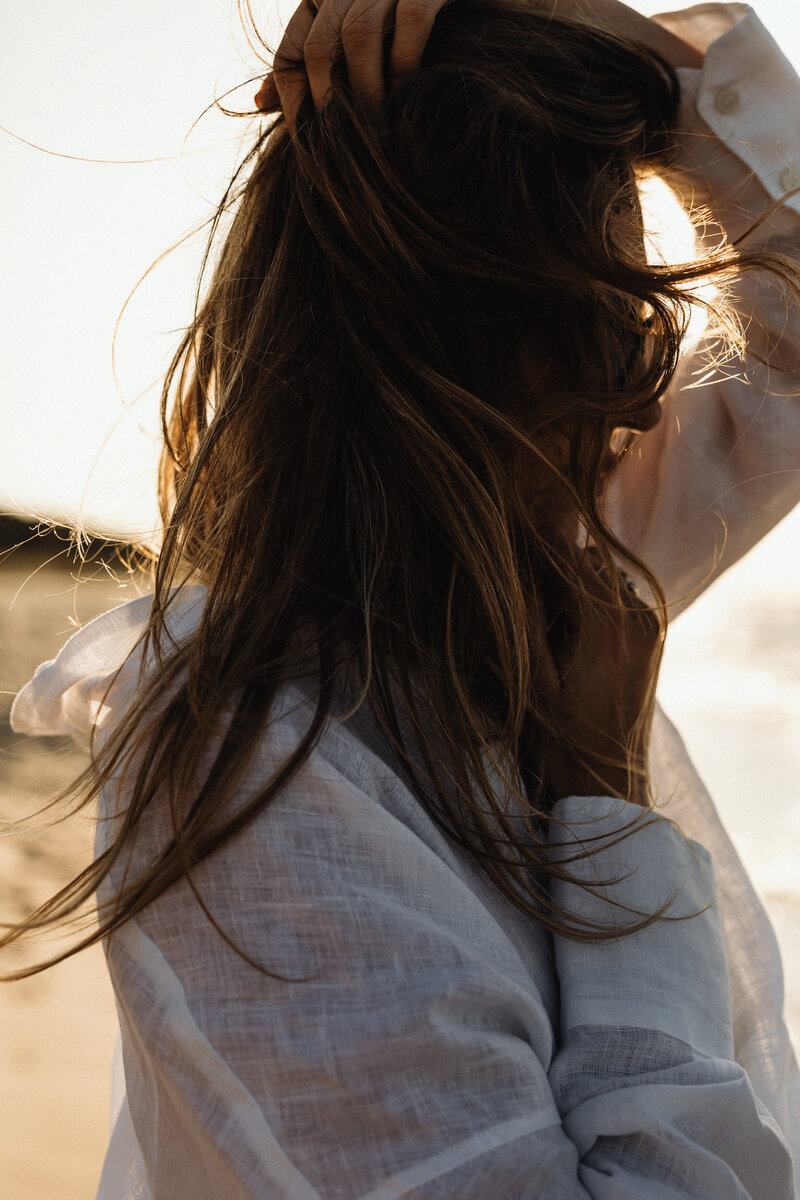 woman on the beach with sunglasses holding her hair from blowing in the breeze at sunset