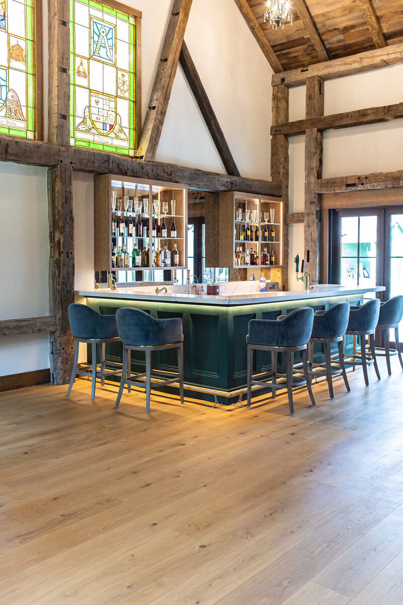 A grand bar room featuring 30-foot ceilings and stunning stained glass windows, reminiscent of cathedral design. The bar is highlighted by large, expensive stools and illuminated from the bottom and floor, creating an opulent and inviting ambiance.