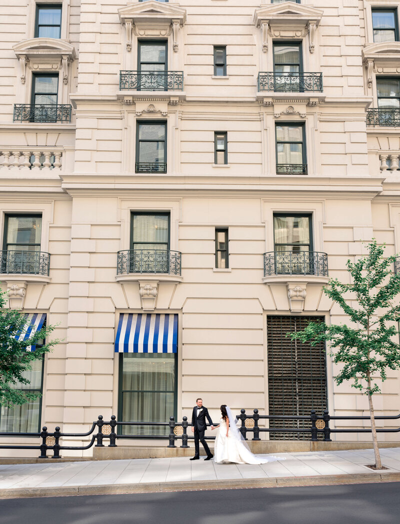A bride in a flowing white gown and a groom in a black suit hold hands, walking in front of an elegant, multi-story building with ornate balconies and striped awnings. The street is lined with black railings and small trees.