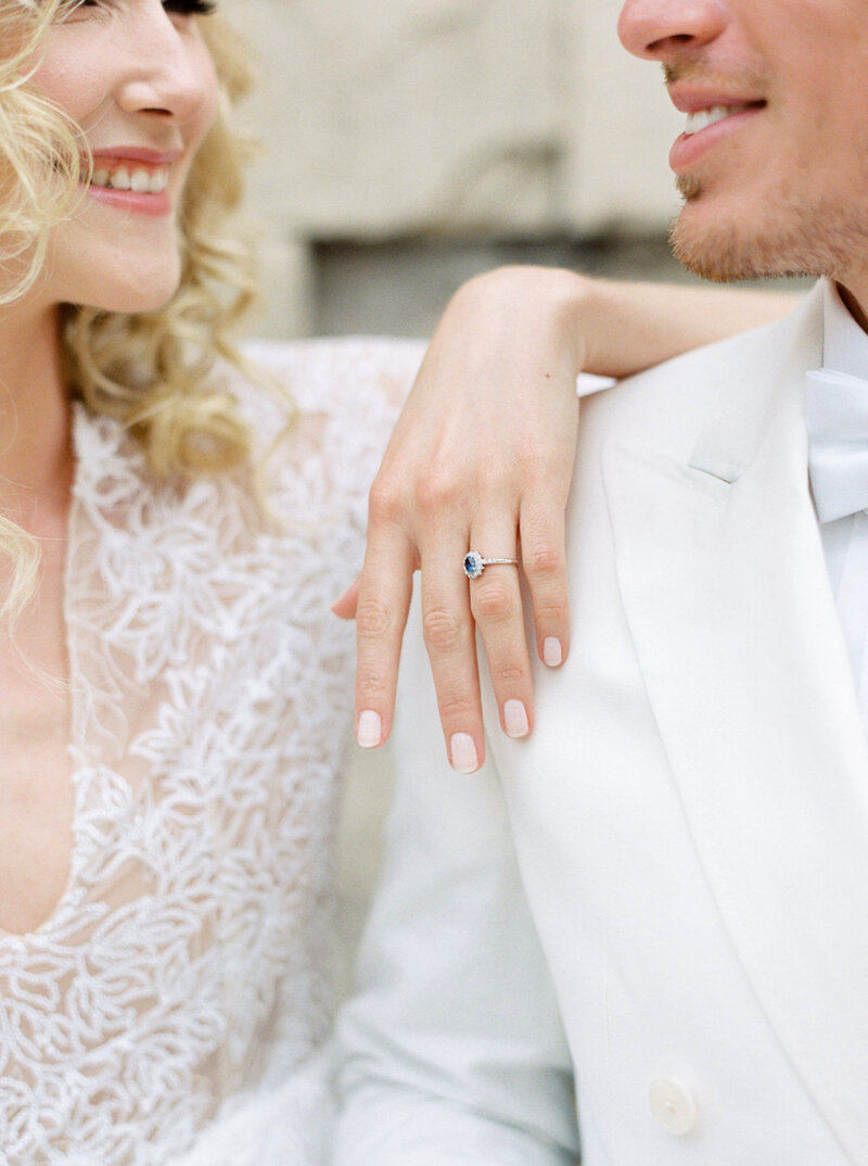 Bride and groom smile at each other while bride rests her arm over grooms shoulder showcasing engagement ring