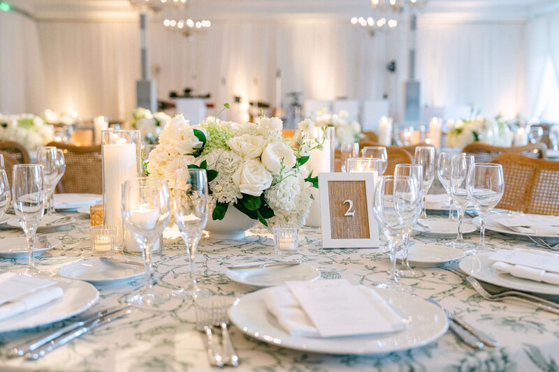 Dior inspired wedding tablescape featuring white hydrangeas  and white roses with greenery in a white bowl vase with rattan and white wood letter pressed table numbers and white candles on the tables with white plates and white napkins and a green and white table cloth