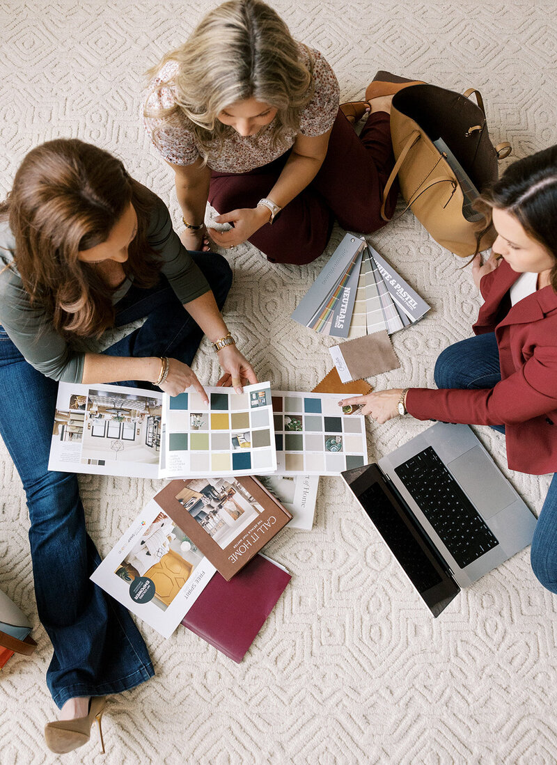 Three women sit on a patterned carpet, reviewing color swatches and design brochures. A laptop is open beside them, and a handbag rests nearby. They appear engaged in a collaborative discussion.