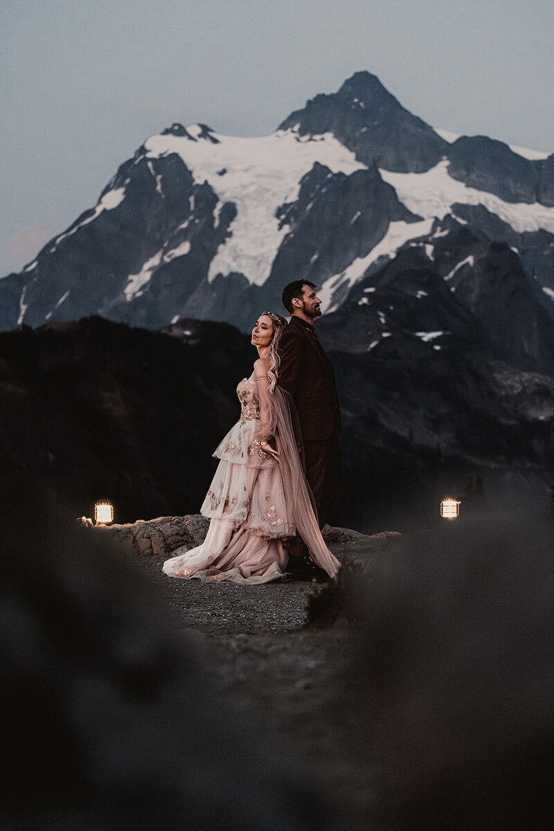a couple stands back to back in their wedding attire just in front of a snowy mountain peak. Their adventure elopement photographers shoot through boulders on the trail to frame the bottom third of the image. 