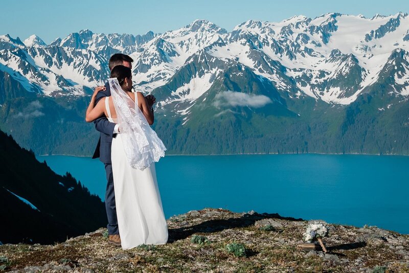 A bride and groom in their wedding finest stand atop a mountain summit of flat rocks covered in tundra grass.  behind them the view drops off to ultra blue water of Resurrection Bay in Seward, Alaska.  There are mountains jagged, snow covered and laced with green shooting up out of the bay, in a spot only accessible via the helicopter they took during their private heli and glamping elopement package.  The bride's long white veil blows in the wind. 