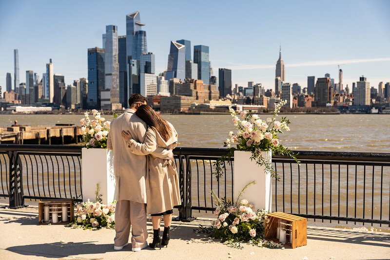 Couple's moment during a romantic proposal setup with floral arrangements overlooking the New York City skyline, styled by Yours Truly, a wedding planner and designer in New Jersey