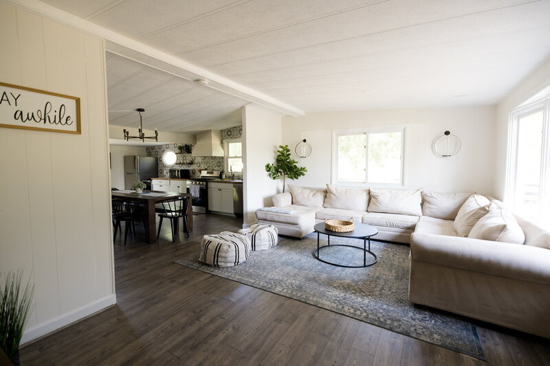 A spacious living room features a large beige sectional sofa, a round coffee table on a textured rug, and two poufs. Sunlight floods through the large windows. In the background, the dining area and kitchen with modern fixtures enhance this inviting venue's charm.