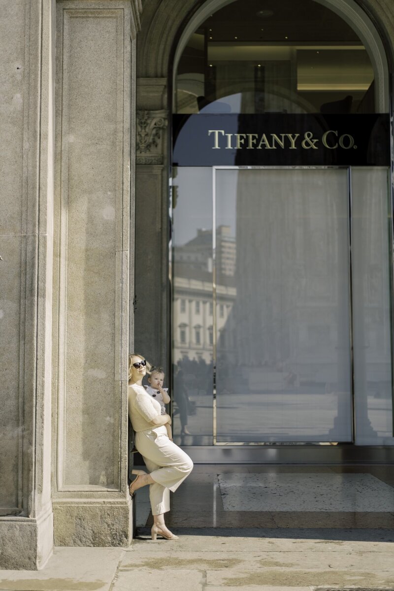 Mother and daughter nestled together outside a boutique, framed by soft architecture and shadow
