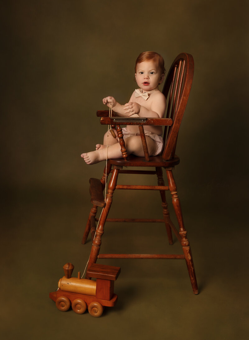 Baby boy poses for his first birthday photoshoot in Brooklyn, NY. He is sitting in an antique wood high chair wearing a diaper cover and bow tie. There is a wooden train on the floor in front of him. He is looking intently at the camera. Captured by premier Brooklyn NY family photographer Chaya Bornstein Photography.