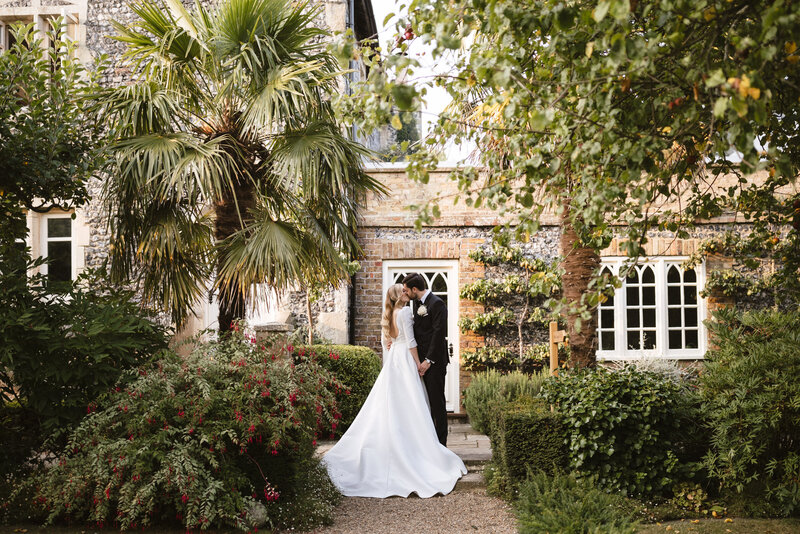 A bride and groom share a romantic moment in a lush garden in front of a charming stone building at Chapel House Estate. The bride wears a flowing white gown and the groom is in a black suit. They are surrounded by greenery and colorful flowers under a sunny sky.