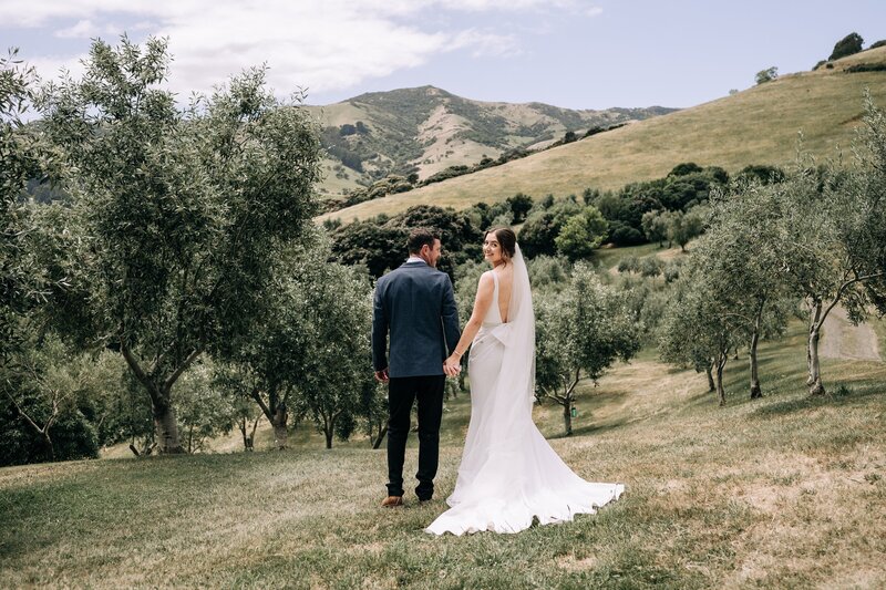 a bride and groom walk through an akaroa olive grove during their wedding photos in akaroa