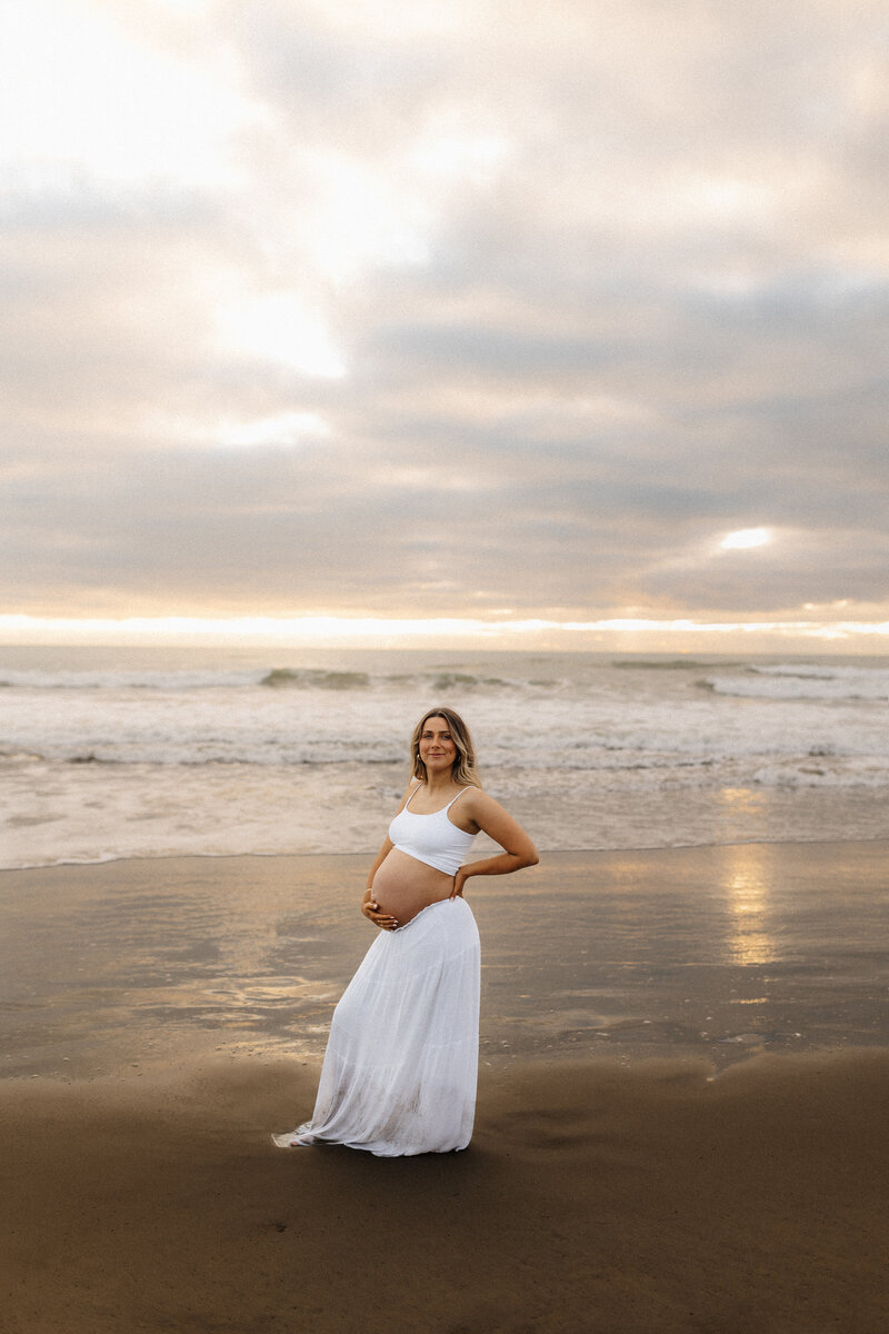 A pregnant woman standing on a sunset beach holding her belly during a maternity photoshoot with christchurch photographer lucy jane
