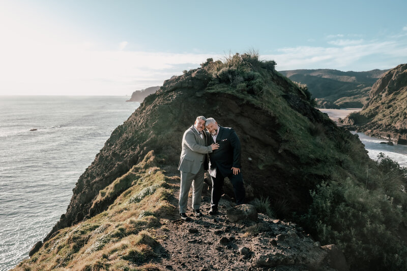 An LGBTQ couple standing on a clifftop holding each other with the ocean view behind them captured by Zanthe Vorsatz Photography