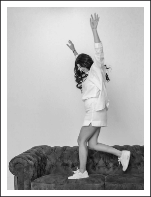 Bel, the founder and owner of Soulve Studio, is captured mid-movement, standing on a sofa with her arms raised, expressing joy and creativity. The image is in black and white, adding a bold artistic flair. A soft purple "S" graphic, representing Soulve Studio, is displayed in the bottom corner, highlighting the studio's brand identity.