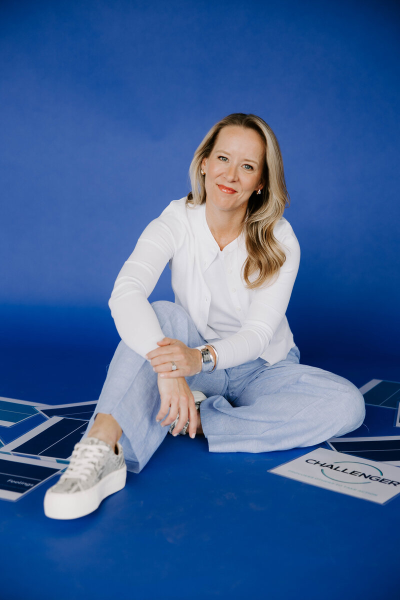 Heather Frick executive coach wearing a white top and jeans sitting on a blue studio background surrounded by coaching papers
