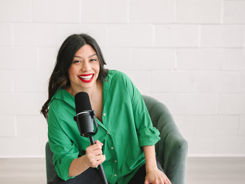 A woman holding a podcast mic and posing in a chair in a studio setting