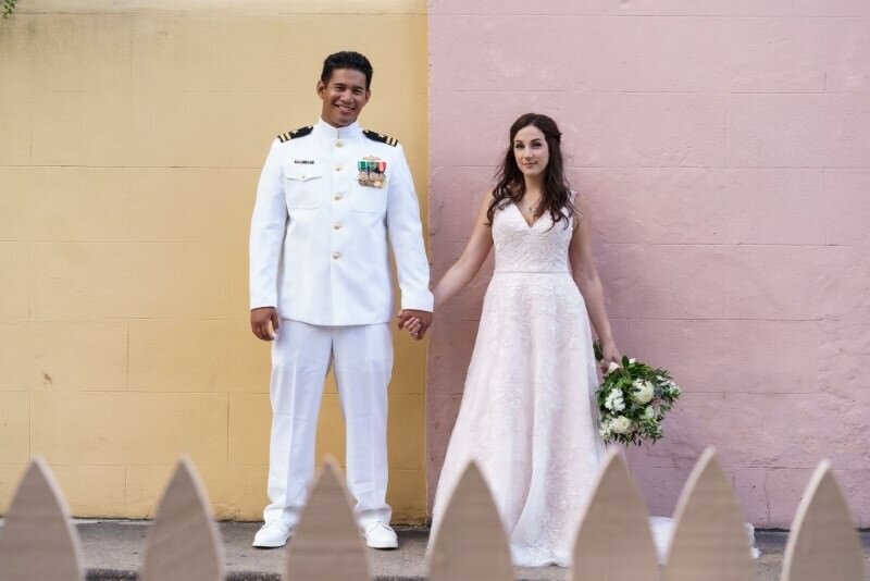 a groom in uniform holds hands with his bride in front of a pink and yellow wall in downtown st augustine