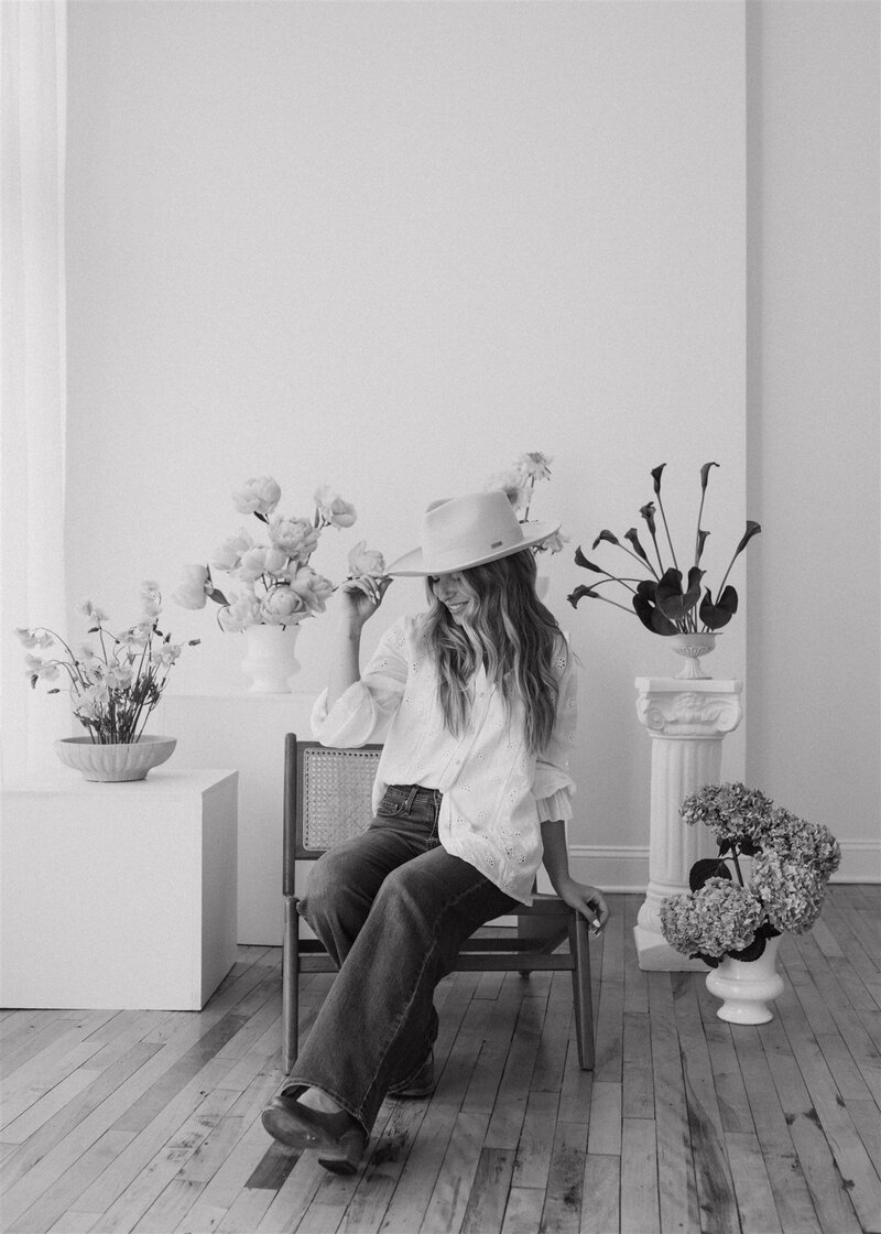 Black and white image of floral designer sitting in a chair surrounded by creative floral arrangements