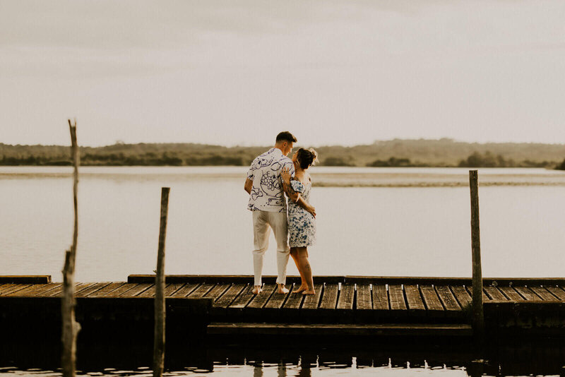 Laura et son futur mari Clément, debout et de dos, s'enlacent tendrement sur un ponton au bord d'un lac.