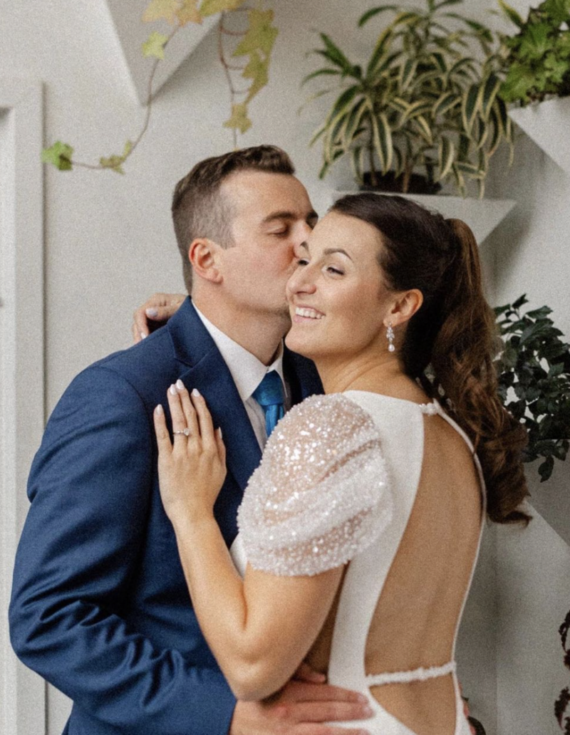 A groom in a blue suit, kissing the bride on the cheek, her hair is in a high ponytail, her dress has sequined shoulders with an open back, her hand is on the groom's chest, ring is on display. Their wedding was coordinated by New Hampshire Wedding Planner - Erika Diedering