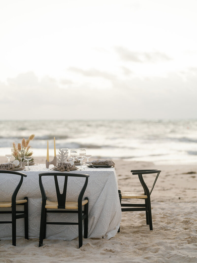 Chairs and a table adorned with light-colored decorations for a wedding reception on a sandy beach in Mexico with water in the background