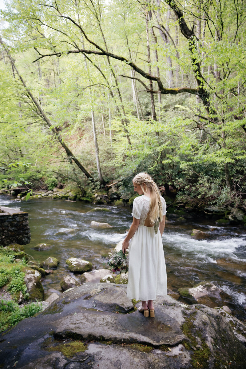 Spence Cabin wedding venue with bride in a lace midi dress holding a bouquet and walking on a rock in the river in the woods near the Gatlinburg wedding venue as she elopes to Gatlinburg 