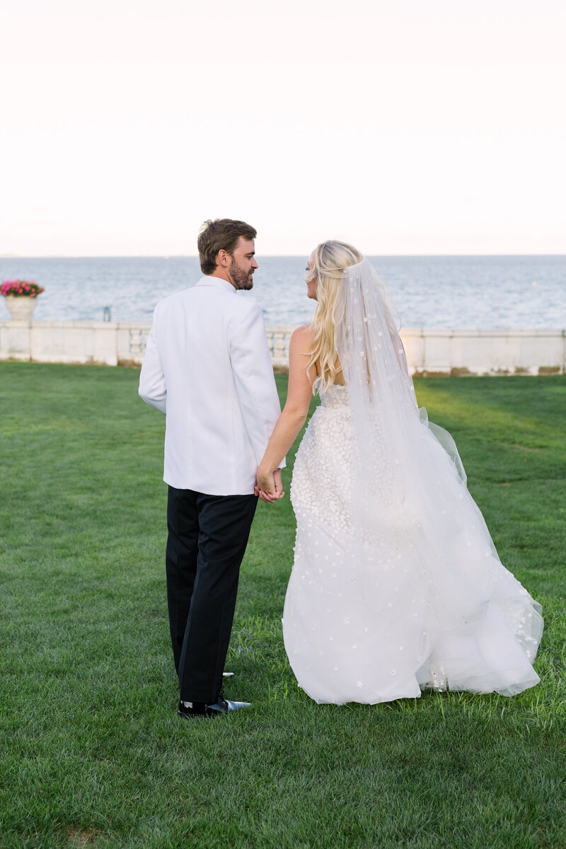 Bride and groom holding hands and walking the grounds at Rosecliff Mansion, overlooking the bay