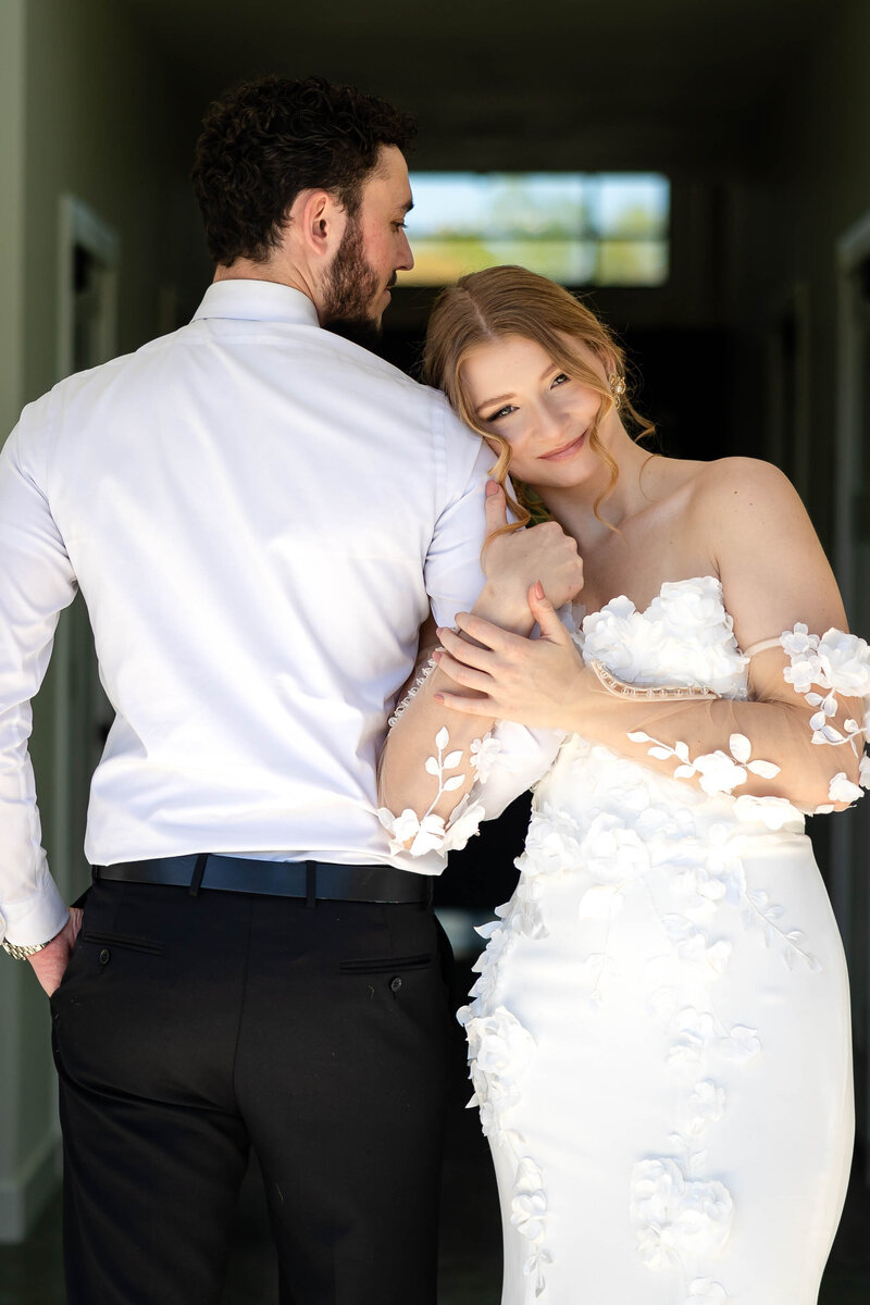Groom with left hand in pocket facing away from the camera but glancing slightly to the right at his bride.  Bride is facing the camera while holding Grooms left arm in an embrace.  Bride has a soft smile and leaning her head against groom shoulder.