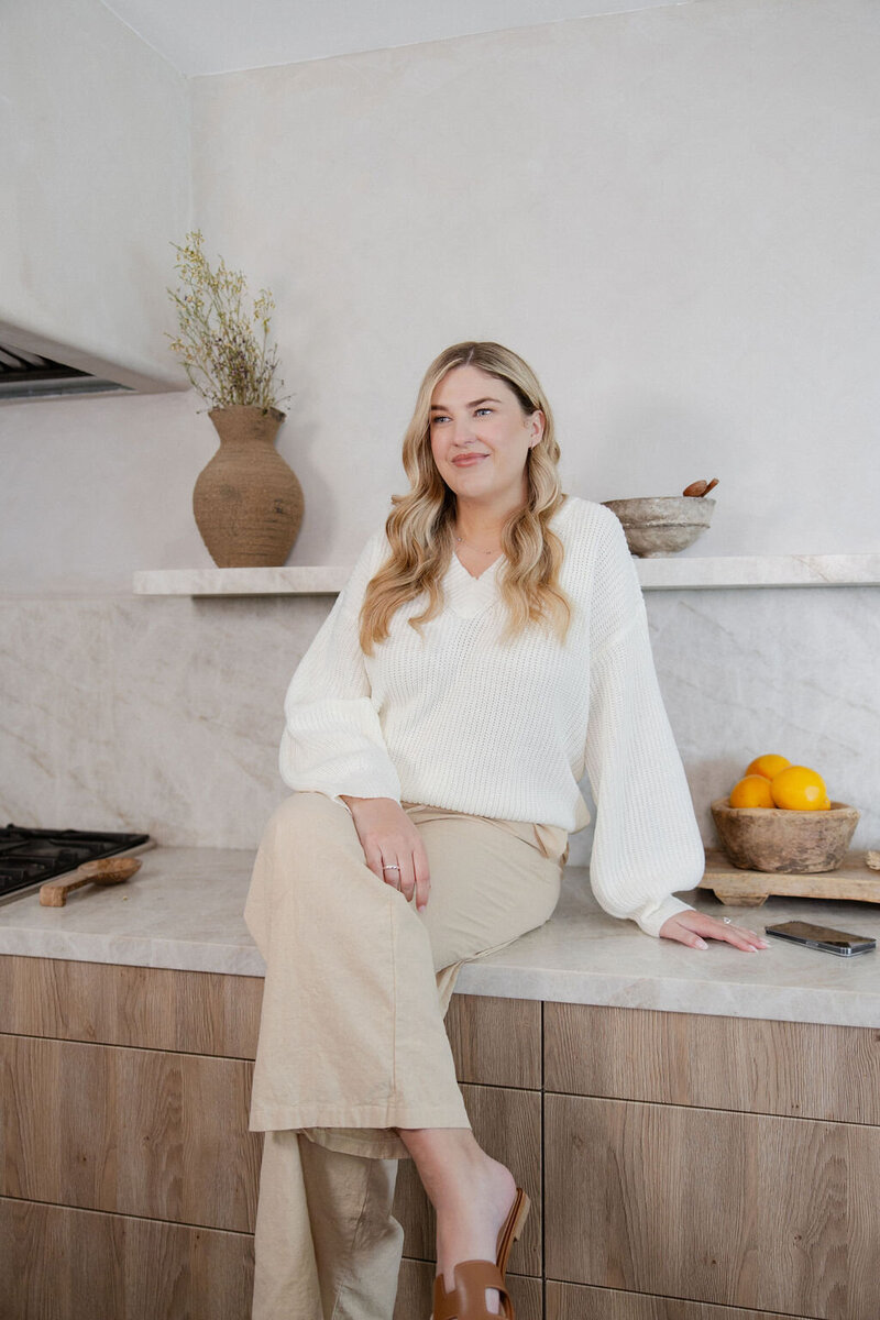 Brogan relaxed and seated on the counter of a modern, neutral kitchen wearing a cozy sweater 