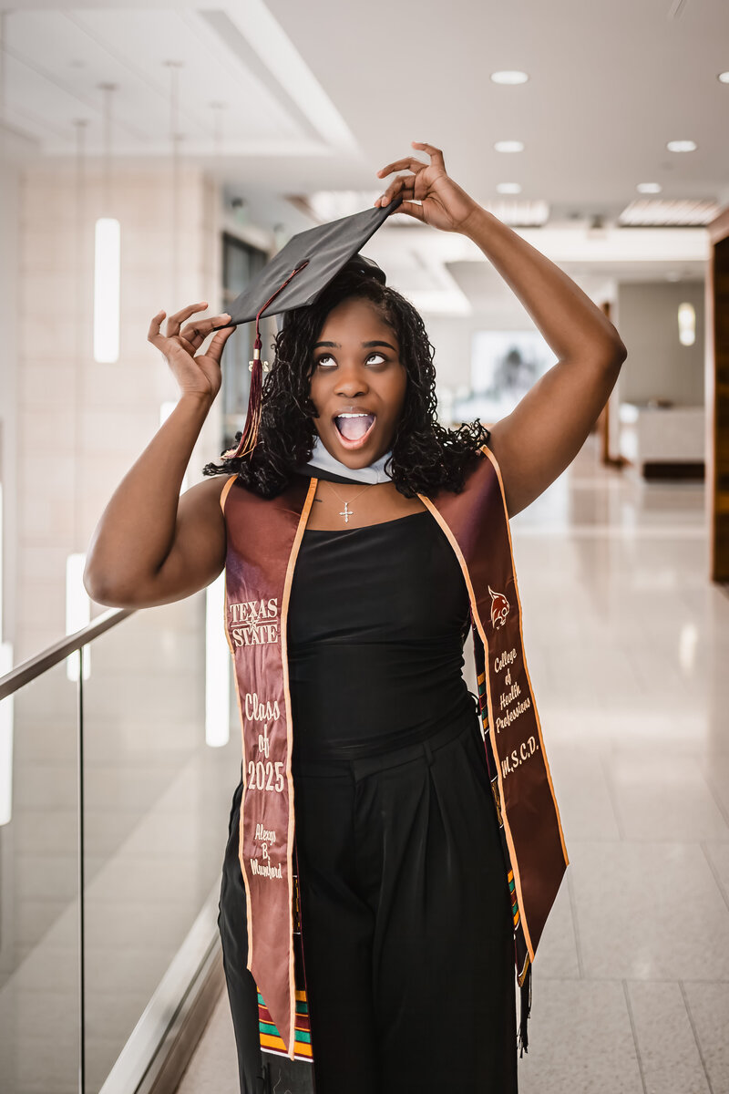 College graduate in black pantsuit at Texas State Campus holding cap on head and smiling.