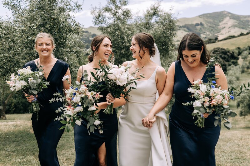 bridesmaids in black and bride laugh as they walk through akaroa olive grove wedding venue