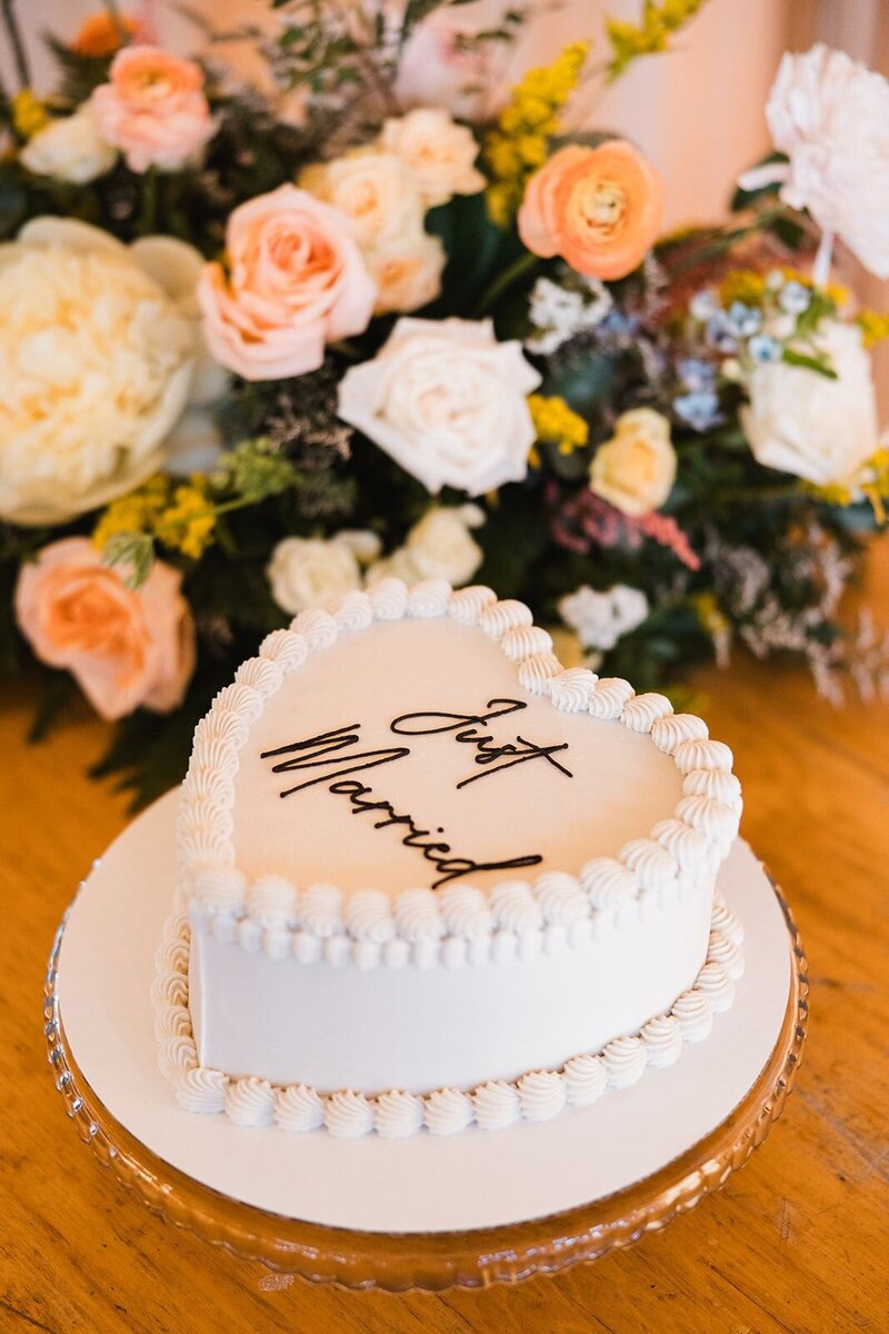 Small white heart shaped cake with just married written on it with flowers in the background