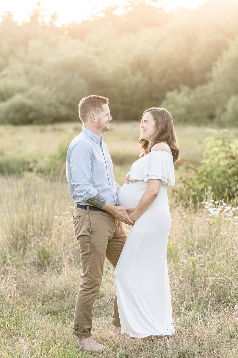 light-skinned man in light blue shirt and khaki pants holding hands and facing pregnant woman dressed in white flowy dress out in nature during a maternity portrait session in nw portland oregon